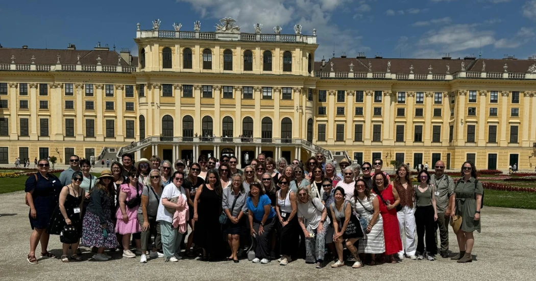 A diverse group of tourists poses in front of a grand Schonbrunn Palace with intricate architectural details under a blue sky.