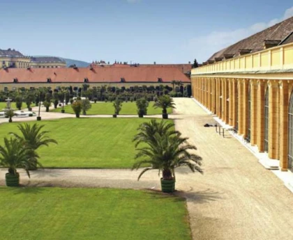The exterior of the Schönbrunn Palace Orangery featuring a grand, baroque façade with large, arched windows, ornate detailing, and a striking green copper roof, set amidst beautiful gardens.