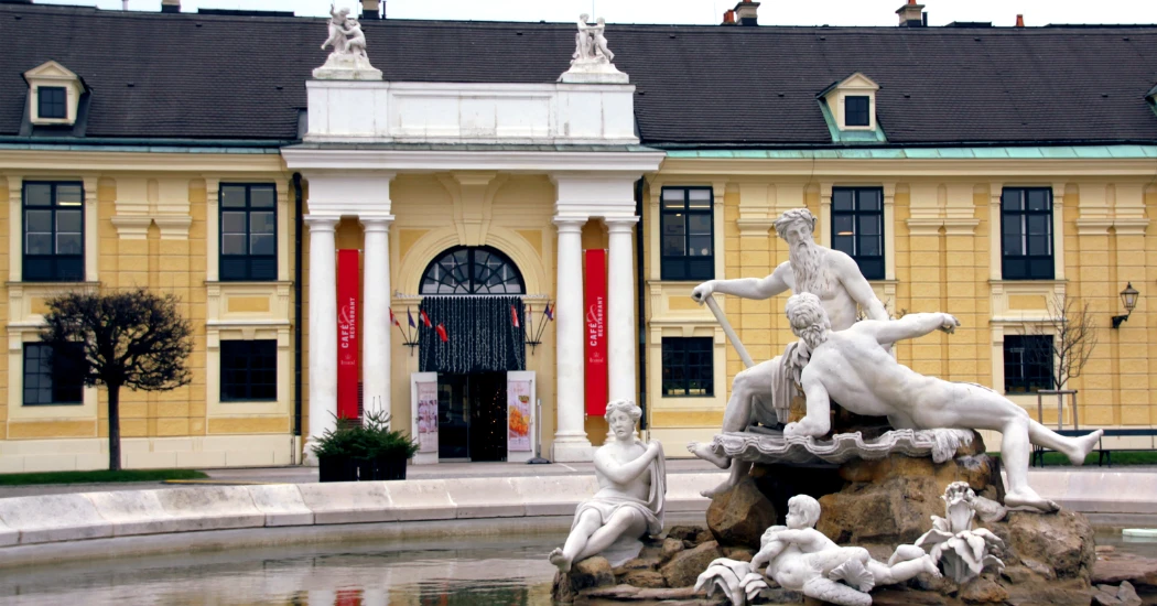 A grand fountain featuring classical statues, set against a historic building with yellow walls and red accents.